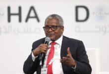 A middle-aged Black man wearing a dark suit, white shirt, and red tie speaks into a handheld microphone while gesturing with his other hand at a conference or public event.