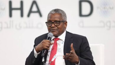A middle-aged Black man wearing a dark suit, white shirt, and red tie speaks into a handheld microphone while gesturing with his other hand at a conference or public event.