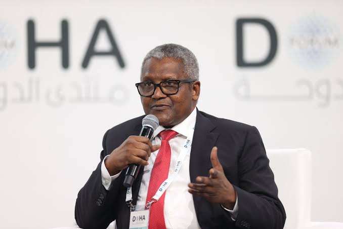 A middle-aged Black man wearing a dark suit, white shirt, and red tie speaks into a handheld microphone while gesturing with his other hand at a conference or public event.