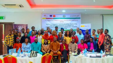 Group photograph of participants at a workshop on peace, security, governance, countering misinformation and disinformation, fact-checking, media literacy, and digital learning. Participants are gathered inside a conference room, with several people seated in front and others standing behind them. A workshop banner is displayed in the background.