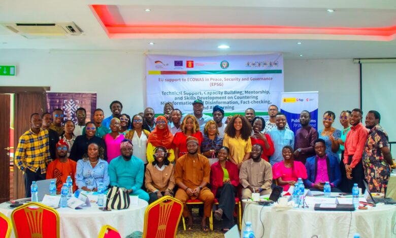 Group photograph of participants at a workshop on peace, security, governance, countering misinformation and disinformation, fact-checking, media literacy, and digital learning. Participants are gathered inside a conference room, with several people seated in front and others standing behind them. A workshop banner is displayed in the background.
