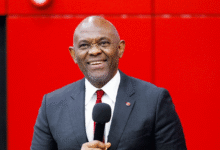 A smiling Black man in a dark gray suit, white shirt, and red tie holds a microphone while standing against a bright red background.