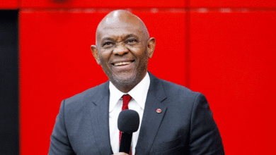 A smiling Black man in a dark gray suit, white shirt, and red tie holds a microphone while standing against a bright red background.