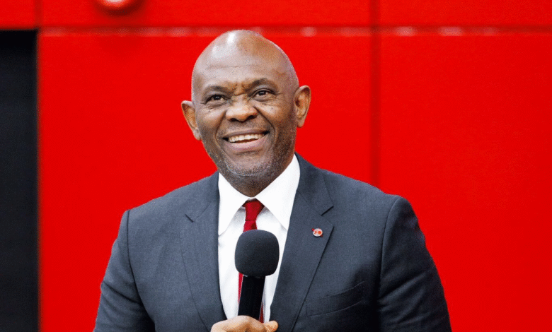 A smiling Black man in a dark gray suit, white shirt, and red tie holds a microphone while standing against a bright red background.