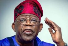 Nigerian President Bola Ahmed Tinubu wearing a red traditional cap and blue attire, raising one hand while speaking.