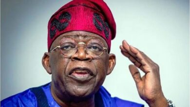 Nigerian President Bola Ahmed Tinubu wearing a red traditional cap and blue attire, raising one hand while speaking.
