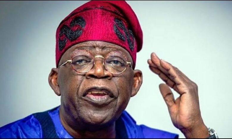 Nigerian President Bola Ahmed Tinubu wearing a red traditional cap and blue attire, raising one hand while speaking.