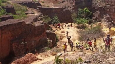A group of people walking and working through a rugged, dry rocky landscape surrounded by steep reddish-brown cliffs and eroded earth.