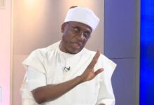 A man wearing a white traditional outfit and cap gestures with one hand while speaking during a televised interview.