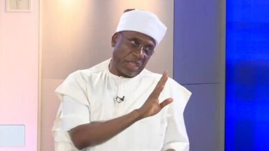 A man wearing a white traditional outfit and cap gestures with one hand while speaking during a televised interview.