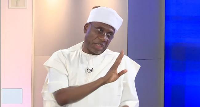 A man wearing a white traditional outfit and cap gestures with one hand while speaking during a televised interview.