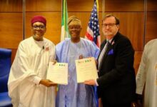 Three men, two Nigerian officials in traditional attire and a US official in a dark suit, pose for a photograph while holding signed documents, with Nigerian and American flags displayed behind them.