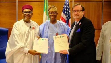 Three men, two Nigerian officials in traditional attire and a US official in a dark suit, pose for a photograph while holding signed documents, with Nigerian and American flags displayed behind them.