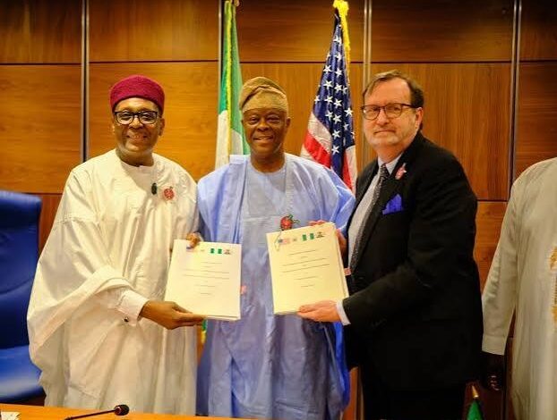 Three men, two Nigerian officials in traditional attire and a US official in a dark suit, pose for a photograph while holding signed documents, with Nigerian and American flags displayed behind them.
