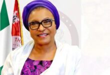 Portrait of a smiling woman wearing a purple headwrap, black-framed glasses, a white jacket and a purple patterned blouse, with flags and an official emblem in the background.