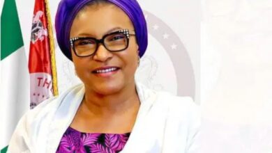 Portrait of a smiling woman wearing a purple headwrap, black-framed glasses, a white jacket and a purple patterned blouse, with flags and an official emblem in the background.