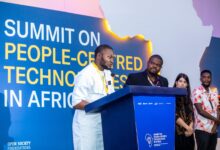 A man in a white traditional outfit speaks at a podium during a summit on people-centred technologies in Africa, with several attendees standing behind him.