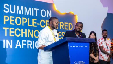 A man in a white traditional outfit speaks at a podium during a summit on people-centred technologies in Africa, with several attendees standing behind him.