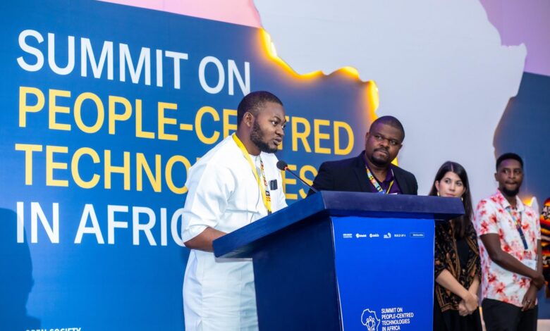 A man in a white traditional outfit speaks at a podium during a summit on people-centred technologies in Africa, with several attendees standing behind him.